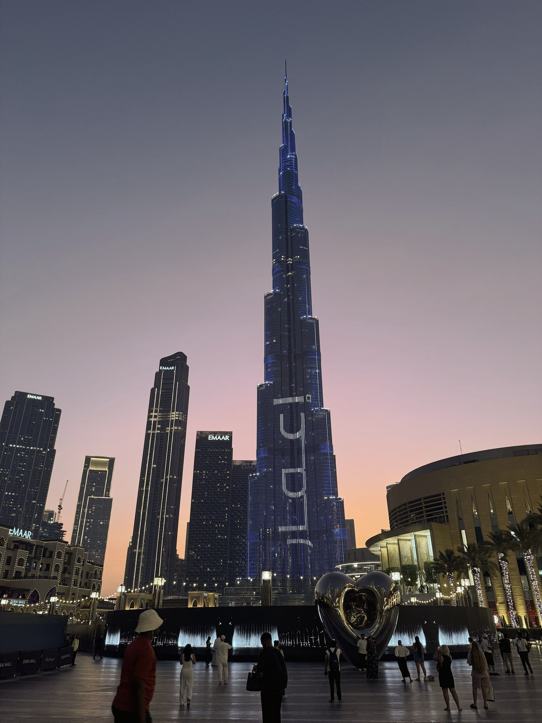 The Burj Khalifa lit up at dusk above Downtown Dubai, with Dubai Mall and the fountain promenade in the foreground.