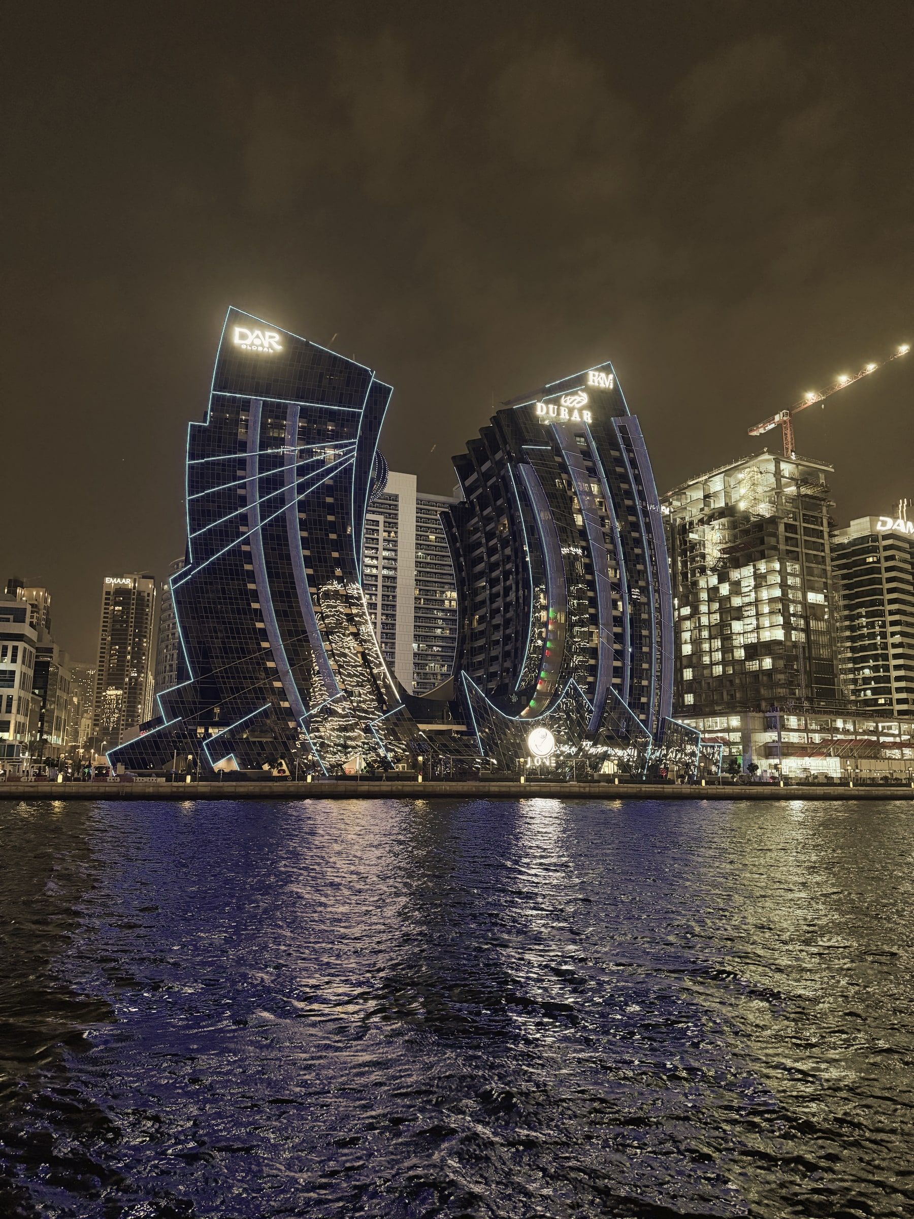 Curved towers outlined in light along the Dubai Water Canal in Business Bay at night, reflected in the water.