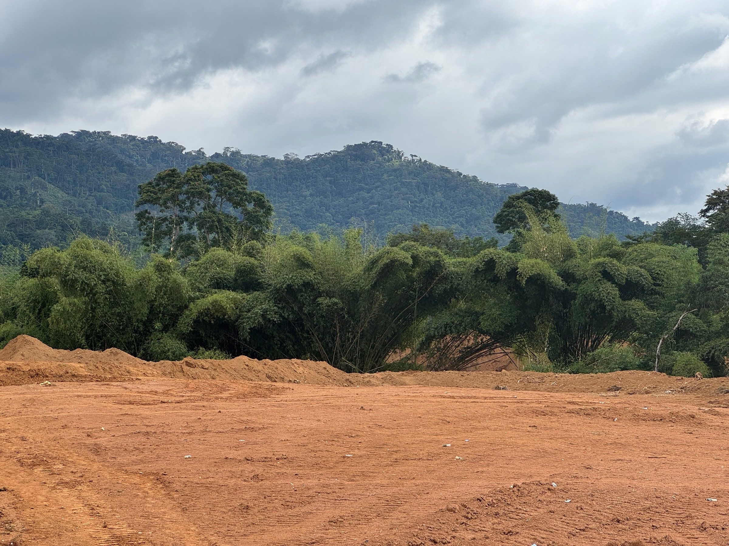 Cleared red earth at a gold sourcing site, with bamboo and forested hills under a cloudy sky.